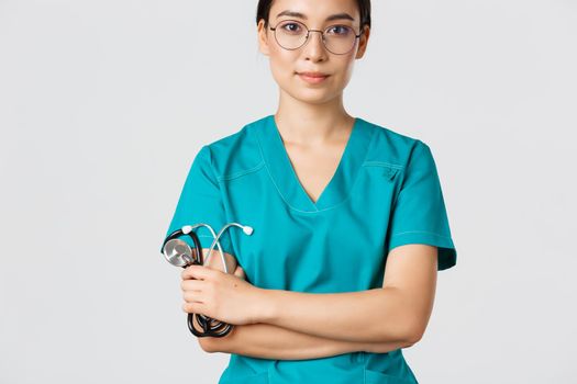 Covid-19, coronavirus disease, healthcare workers concept. Confident smiling professional asian doctor in glasses, cross arms chest, wearing scrubs and holding stethoscope, white background