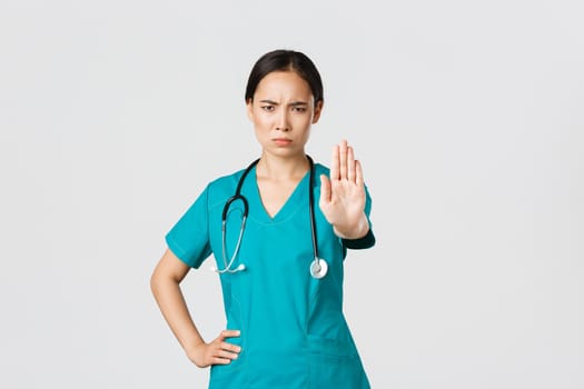 Covid-19, healthcare workers, pandemic concept. Angry serious-looking asian doctor, female physician or nurse in scrubs frowning displeased, extend hand to show stop, disagree, prohibit or forbid