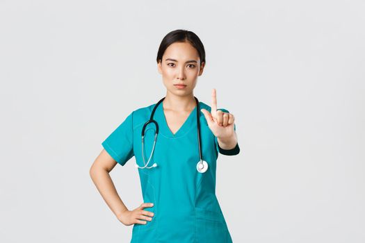 Covid-19, healthcare workers, pandemic concept. Serious-looking professional female asian doctor in scrubs, physician shaking finger in prohibition, warning gesture, forbid something