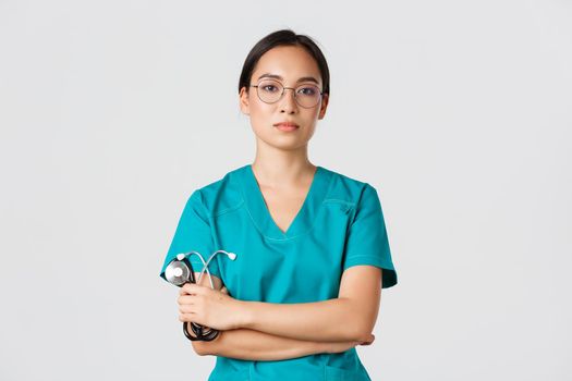 Covid-19, coronavirus disease, healthcare workers concept. Professional confident, serious-looking female doctor, physician in glasses and scrubs, holding stethoscope, standing white background