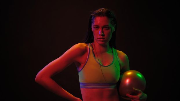 Athlete girl with power exercise ball standing in neon lights wearing sport top working on a weight fitness isolated on black background. Neon light concept.