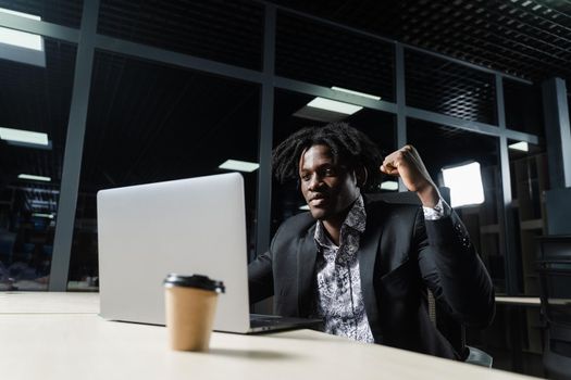 Happy black man with clenched fists is looking on the screen of laptop and rejoice finishing his job.