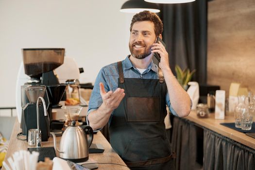 Barista with stylish beard smiling and talk phone on his workplace at coffeehouse
