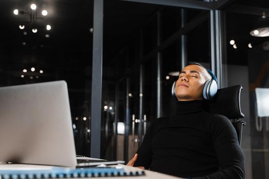 Overtime working. Asian man with closed eyes is resting at work at laptop. Tired male programmer taking a break due to overtime work