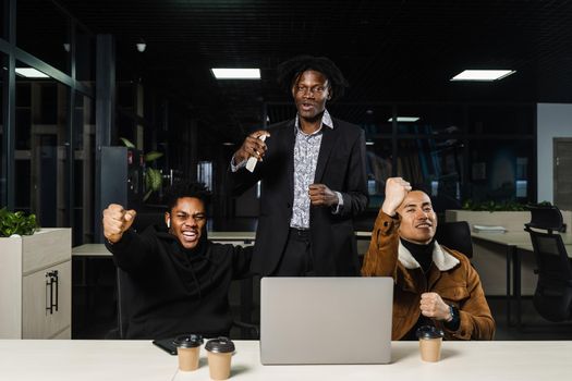 Emotional and joyful black Africans and Asians teamwork together and rejoice at end of online project. Multiethnic colleagues working on laptop, raising their hands, fists and shouting loudly.