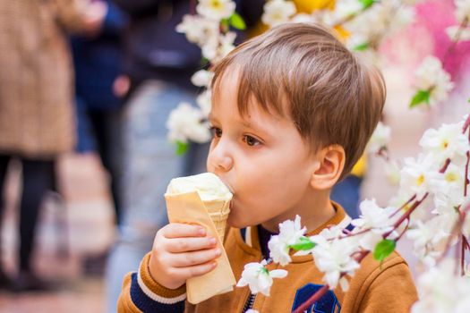 A happy child eats ice cream. A smiling little boy holds 1 ice cream in his hands, hidden by the flowering branches of a tree.