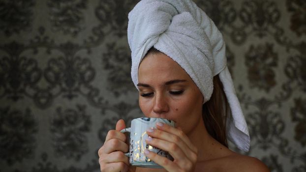 Middle-aged woman looks good with bare shoulders in a white towel on her head holds a cup and drinks coffee or tea against the wall