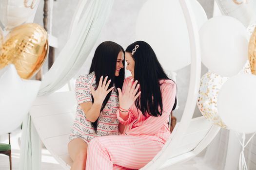 Two happy female friend in pajamas sitting on stylish armchair at luxury interior