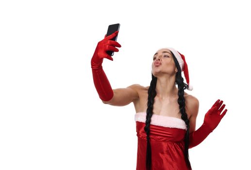 Attractive sexy dark-haired young woman with two pigtails sending kisses while taking a selfie, looking into the camera on a smartphone in her outstretched hand. Copy ad space. Christmas concept