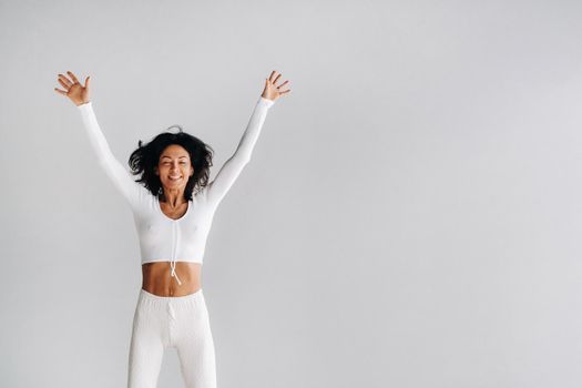 A happy woman in white sportswear bounces on a white background. The girl, jumping, raised her hands up in the gym.