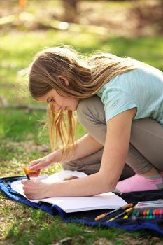A young adolescent girl writes or colors in a notebook or journal on a blanket on the grass