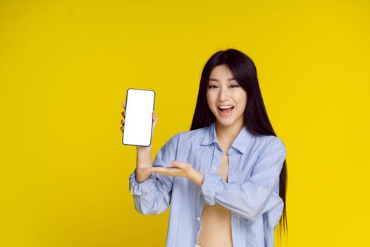 Young happy asian girl holding smartphone showing a white empty screen and exciting to win isolated on yellow background. Great offer. Product placement. Mobile app advertisement