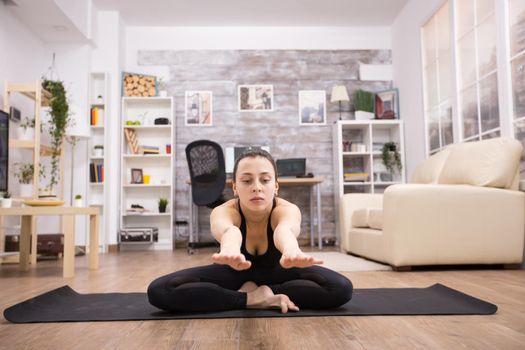 Young woman in sportwear sitting on lotus yoga pose stretching forward in home.