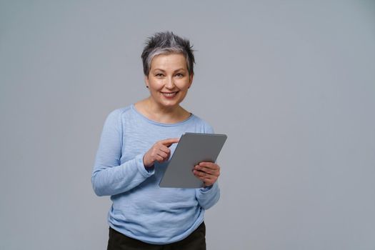 Smiling mature grey haired woman 50s holding digital tablet working or checking on social media. Pretty woman in 50s in blue blouse isolated on white. Older people and technologies. Toned image