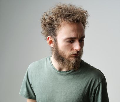 Pensive young handsome bearded man with wild curly hair, bright blue eyes looking down isolated on white background. Young thinking man in green t shirt on white