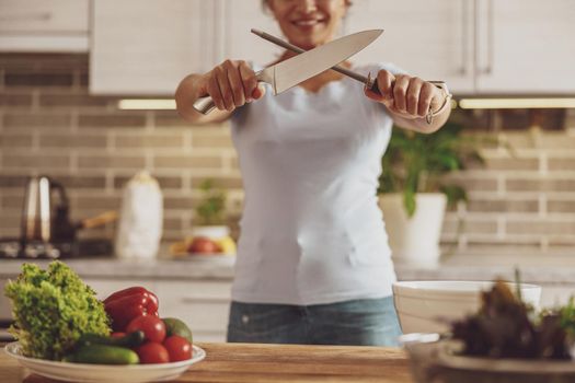 A housewife sharpens a knife standing at a table against the background of the kitchen