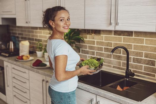 A young woman stands in front of a sink with a plate of greens in her hands.
