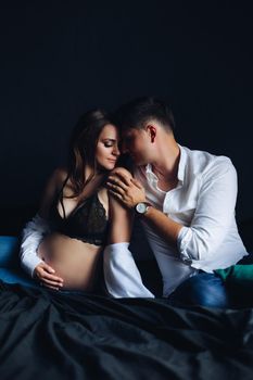 Studio portrait of beautiful and tender couple on bed. Attractive young pregnant woman in black laced bra with loving husband tenderly touching her shoulder. They are sitting face to face over black background.