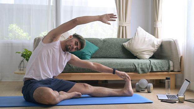 Young man in sports clothes is warming up, stretching while sitting on mat at home