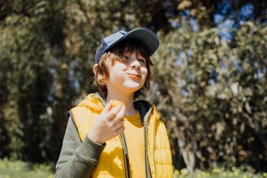 Smiling cheerful child kid in yellow vest and green hoodie eats crisp snacks outdoors in public park. Schoolboy boy enjoying consumes chews junk food outside with trees vegetation on the background