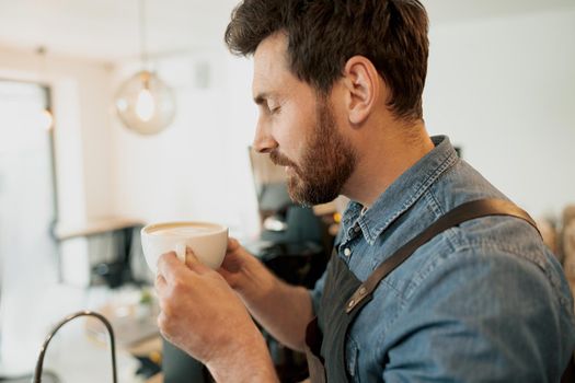 Handsome barista with stylish beard smelling coffee in cafeteria