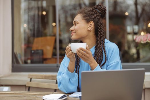 Dreamy young mixed-race stylish female with braids holding cup of coffee at laptop and dreaming.