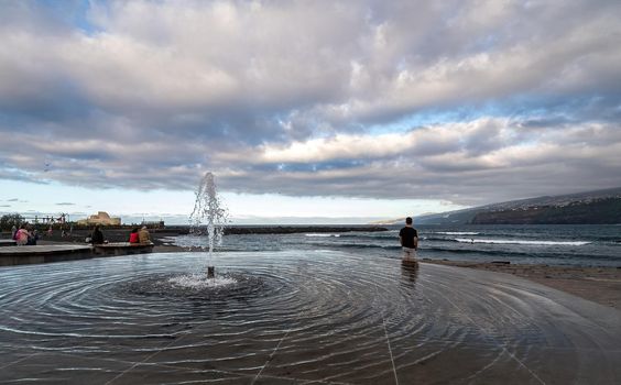 Fountain with water round ripples and a man looking at sea with surfers.