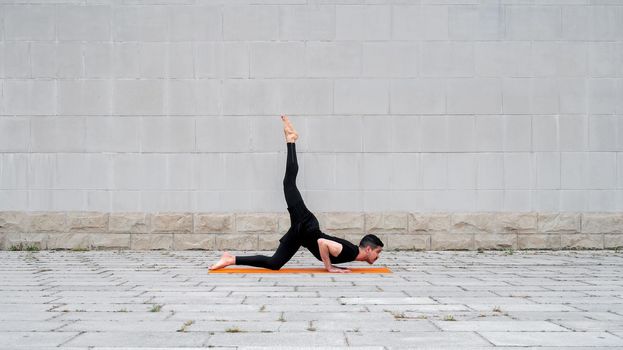 Chaturanga pose with one leg up. Fit latin man do yoga outdoor on orange mat with gray concrete wall at the background.