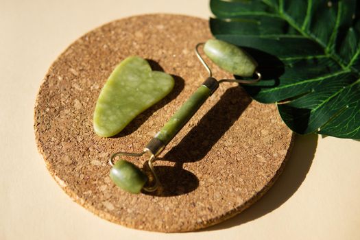 Jade Gua sha scraper and face roller massager on a cork round stand with a monstera leaf. Hard light, shadows, the concept self-care. Facial care. Zero waste. Lifting and toning treatment at home.