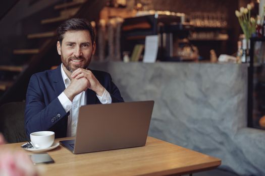 Attractive young elegant businessman having coffee at laptop in cafe and smiling. Chin on hands.