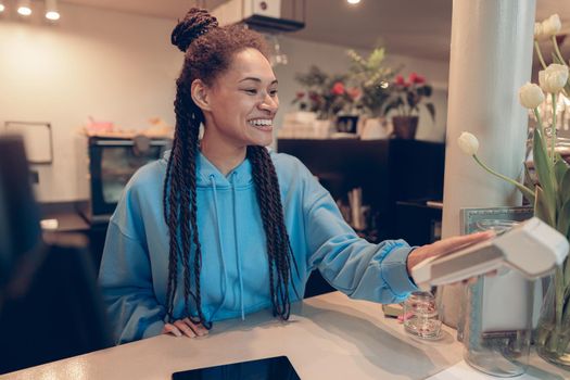 Joyful young mixed-race woman bartender handing payment terminal for client in cafe at counter.