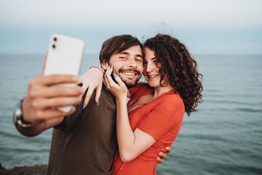 Happy Caucasian Couple Man and Curly Brunette Woman Hugging While Standing Against the Sea and Making Selfie on Smartphone