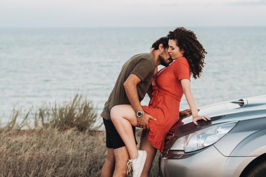 Young Couple on Road Trip, Man Intimate Kissing Woman in the Neck on the Hood of Their SUV Car on Background of the Sea at Sunset