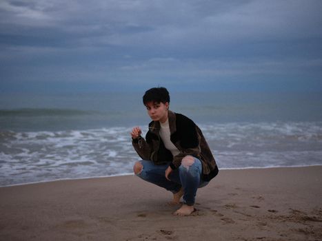 trans woman squatting with the beach behind her, posing in warm clothes