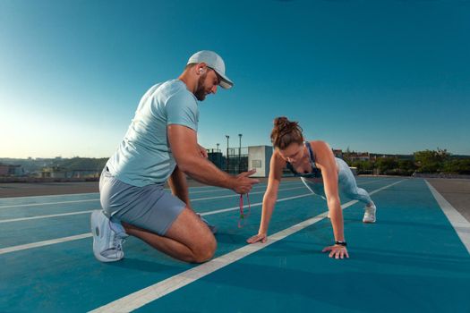 Instructor and athlete runner on the track. The athletics marks the time in the plank exercise on a stopwatch. Fitness trainer and mentee.