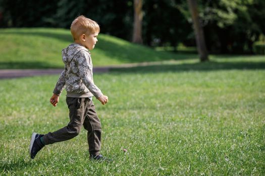 Cute boy running across grass und smiling. An agile happy child is playing sports in a summer city park.