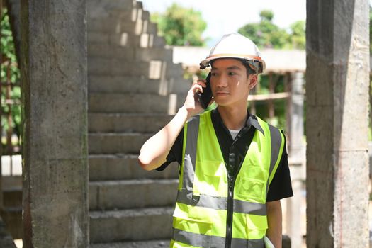 Young engineer builder in helmet talking on mobile phone while standing at construction site