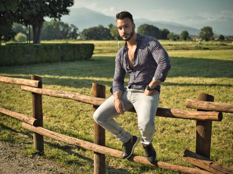 Stylish man sitting on fence in countryside