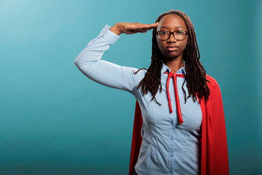 Serious and confident superhero woman standing on blue background while saluting patriotic.