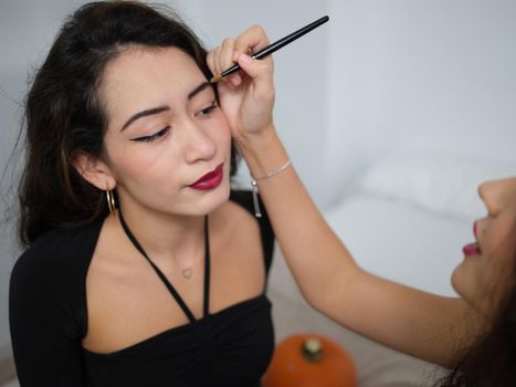 A young woman doing makeup eyebrow pencil to her female friend on halloween