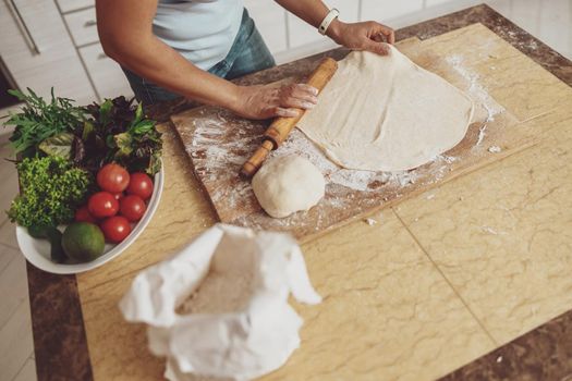 Cooked pizza dough a woman rolls out on a wooden board on the kitchen table