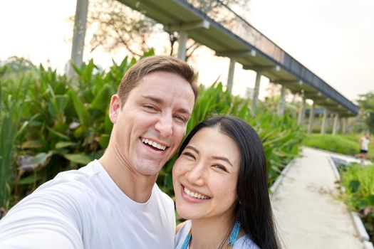 Multiethnic couple laughing while taking a selfie in an urban park during sunset