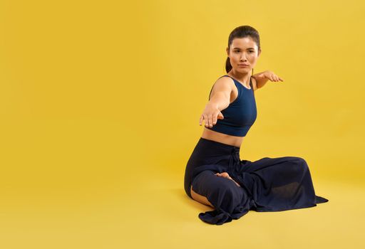 Serious girl holding outstretched arm in front of camera, while sitting in yoga pose indoor. Portrait view of lady with distant look, practicing yoga, isolated on orange background. Concept of yoga.