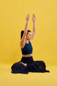 Barefoot girl practicing yoga, with closed eyes alone on floor. Side view of woman in black sportswear sitting in lotus asana with gazed up hands, isolated on studio background. Concept of meditation.