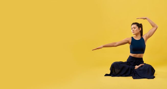 Sporty female performing yoga asana in studio. Front view of sporty woman sitting with raised hand over head while exercising, isolated on yellow studio background, copy space. Concept of yoga asana.
