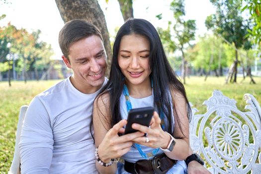 Asian woman and man using the mobile while sitting together on a bench in a park