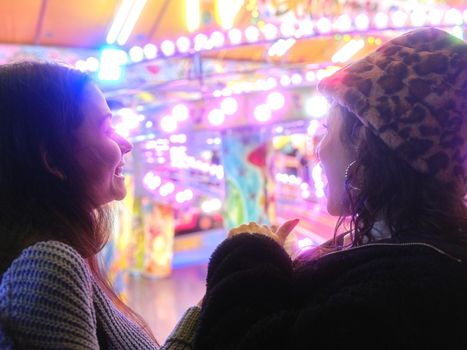 Two women talking in front of an attraction of a colorful evening fair