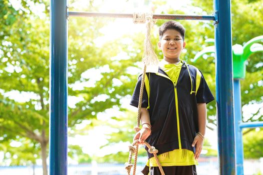 Young asian boy hang the yellow bar by his hand to exercise at out door playground under the big tree.
