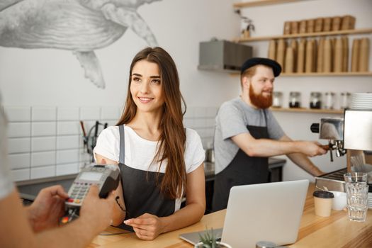 Coffee Business Concept - Beautiful female barista giving payment service for customer with credit card and smiling while working at the bar counter in modern coffee shop.