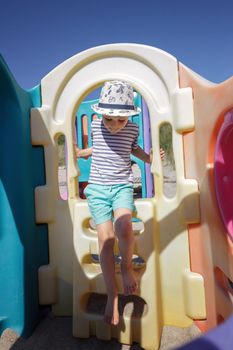 A little boy with a hat climbs a staircase on a colorful plastic playground and performs coordination exercises.